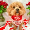A curly-haired dog wears a red and white “Hug Me” bandana, sitting between two vases of red roses on a white backdrop, with scattered rose petals on the floor.