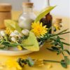 A plate with yellow dandelions, green leaves, and white flower buds sits on a yellow table. In the background are glass jars and bottles, some with cork tops, and a wooden container.