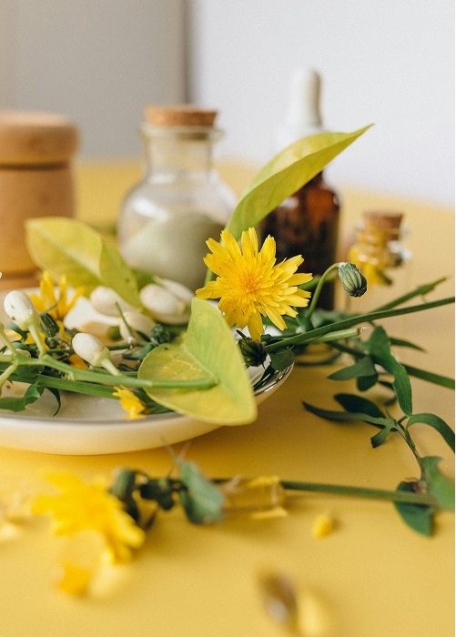 A plate with yellow dandelions, green leaves, and white flower buds sits on a yellow table. In the background are glass jars and bottles, some with cork tops, and a wooden container.
